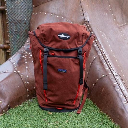 The Patagonia Brown Fieldsmith Lid Pack, featuring black straps and a fish-shaped logo, stands upright on artificial grass in front of a metal slide.