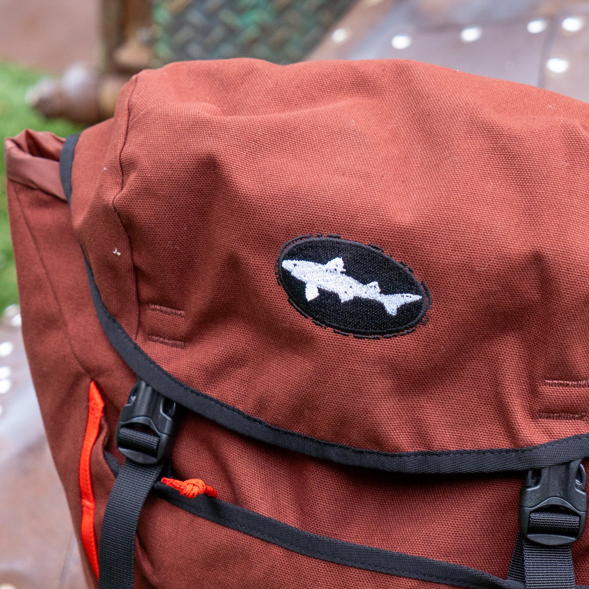 Close-up of the Patagonia Brown Fieldsmith Lid Pack, a rust-colored backpack with black trim, featuring a black oval patch with a white shark silhouette on the top flap.