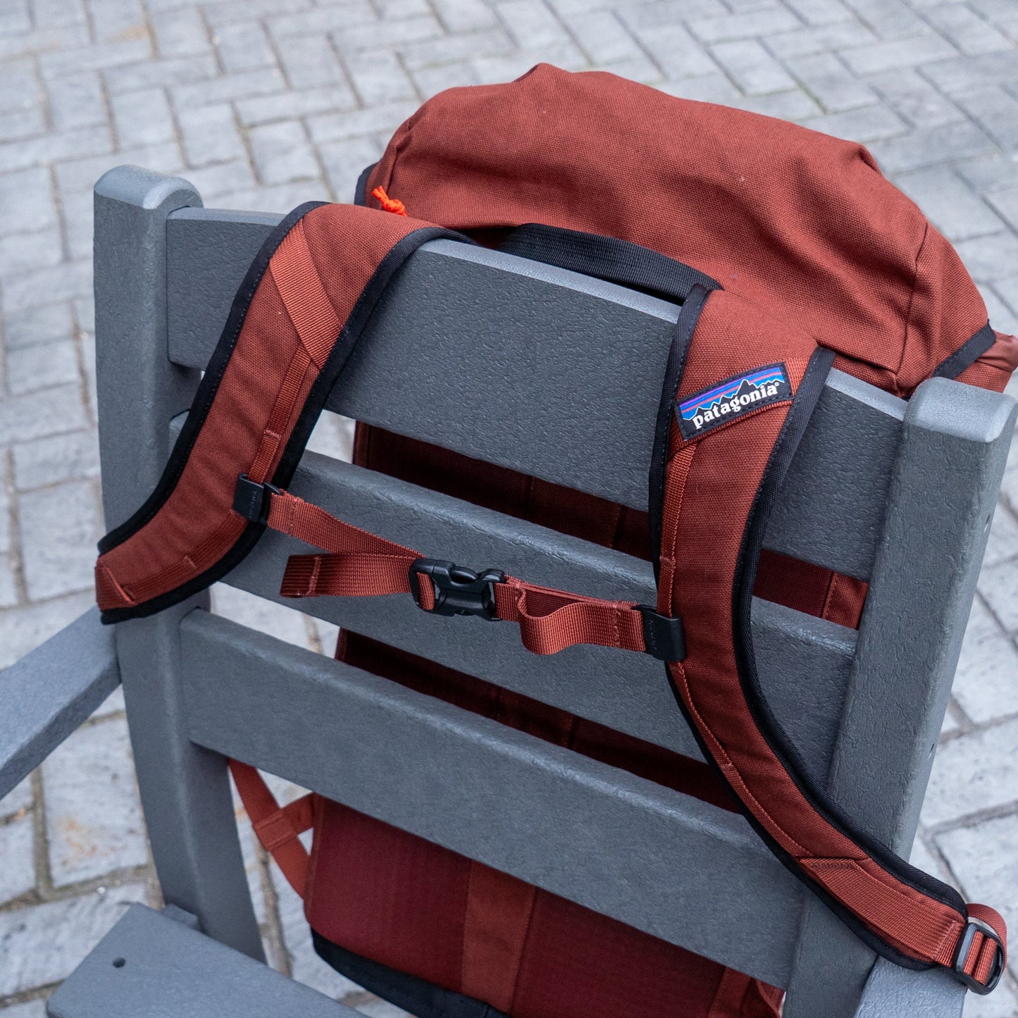 A Patagonia Brown Fieldsmith Lid Pack hangs from a gray outdoor chair on a stone patio, with its shoulder straps and chest buckle visible.