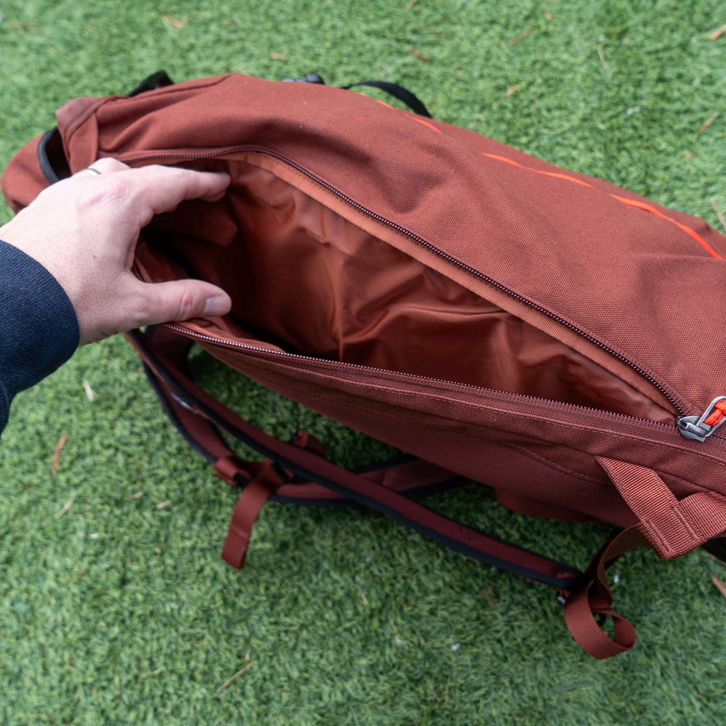 Someone opens the main compartment of a Patagonia Brown Fieldsmith Lid Pack backpack, showing its empty interior while standing on green grass.