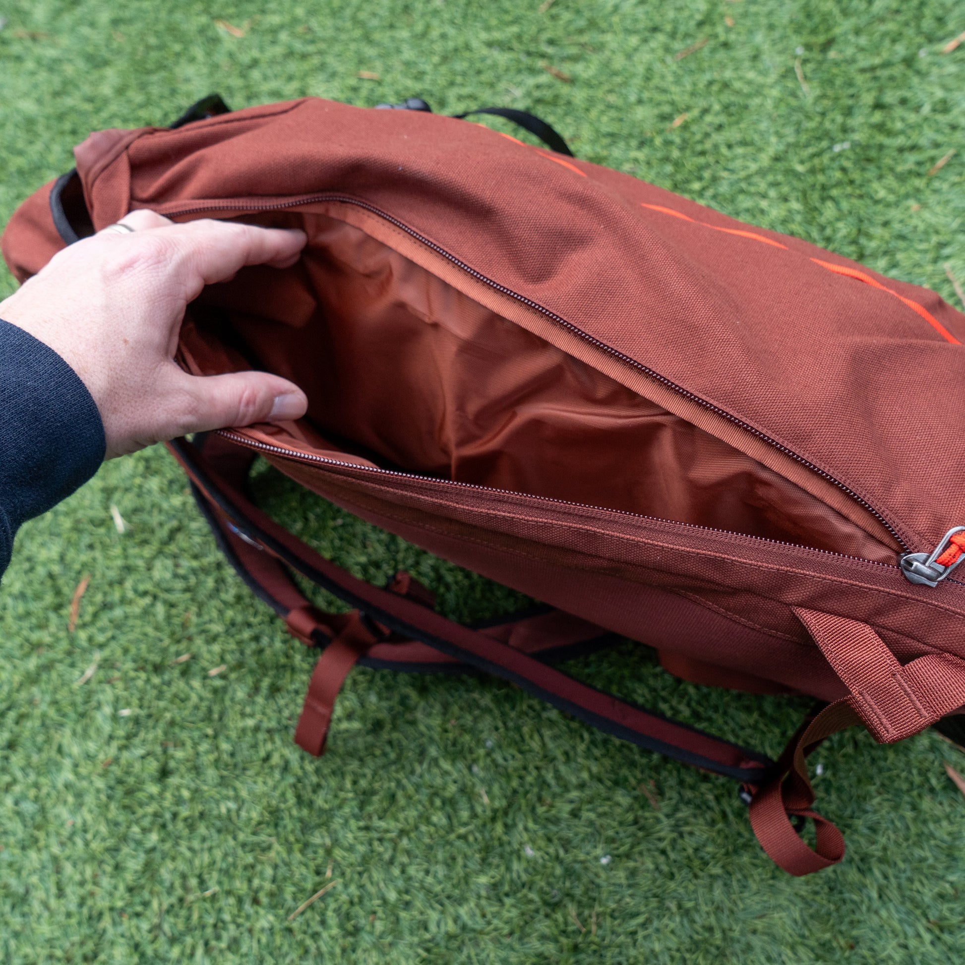 Someone opens the main compartment of a Patagonia Brown Fieldsmith Lid Pack backpack, showing its empty interior while standing on green grass.