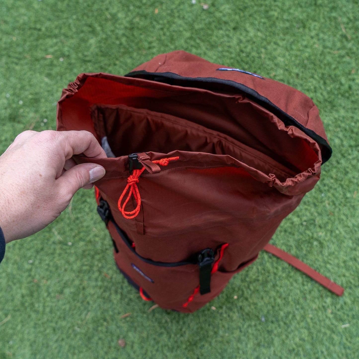 A hand opens the top drawstring closure of a Patagonia Brown Fieldsmith Lid Pack, which stands upright on green grass.