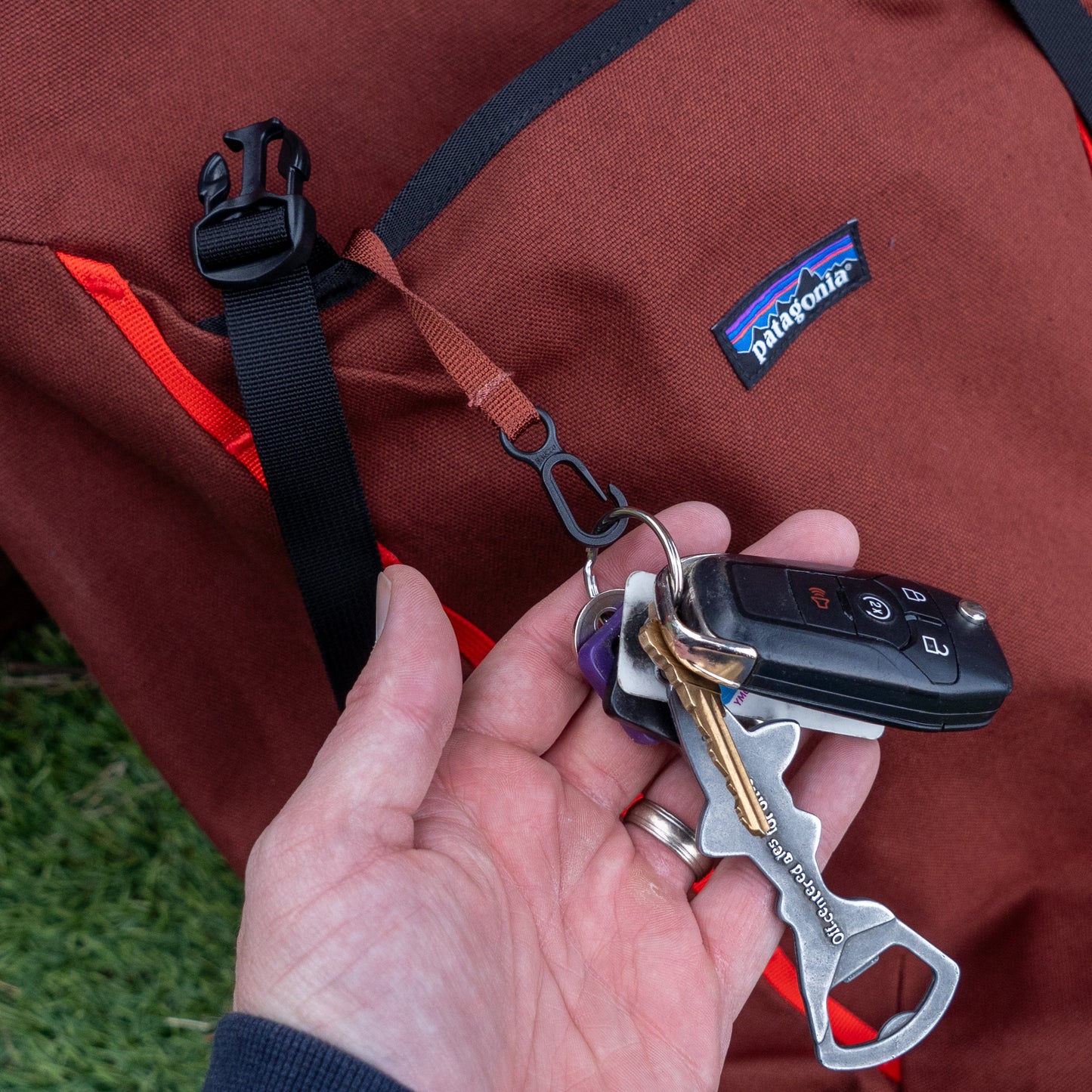A hand holds keys—including a car key, house key, and metal multitool—attached to a Patagonia Brown Fieldsmith Lid Pack with a red strap. Grass is visible in the background.