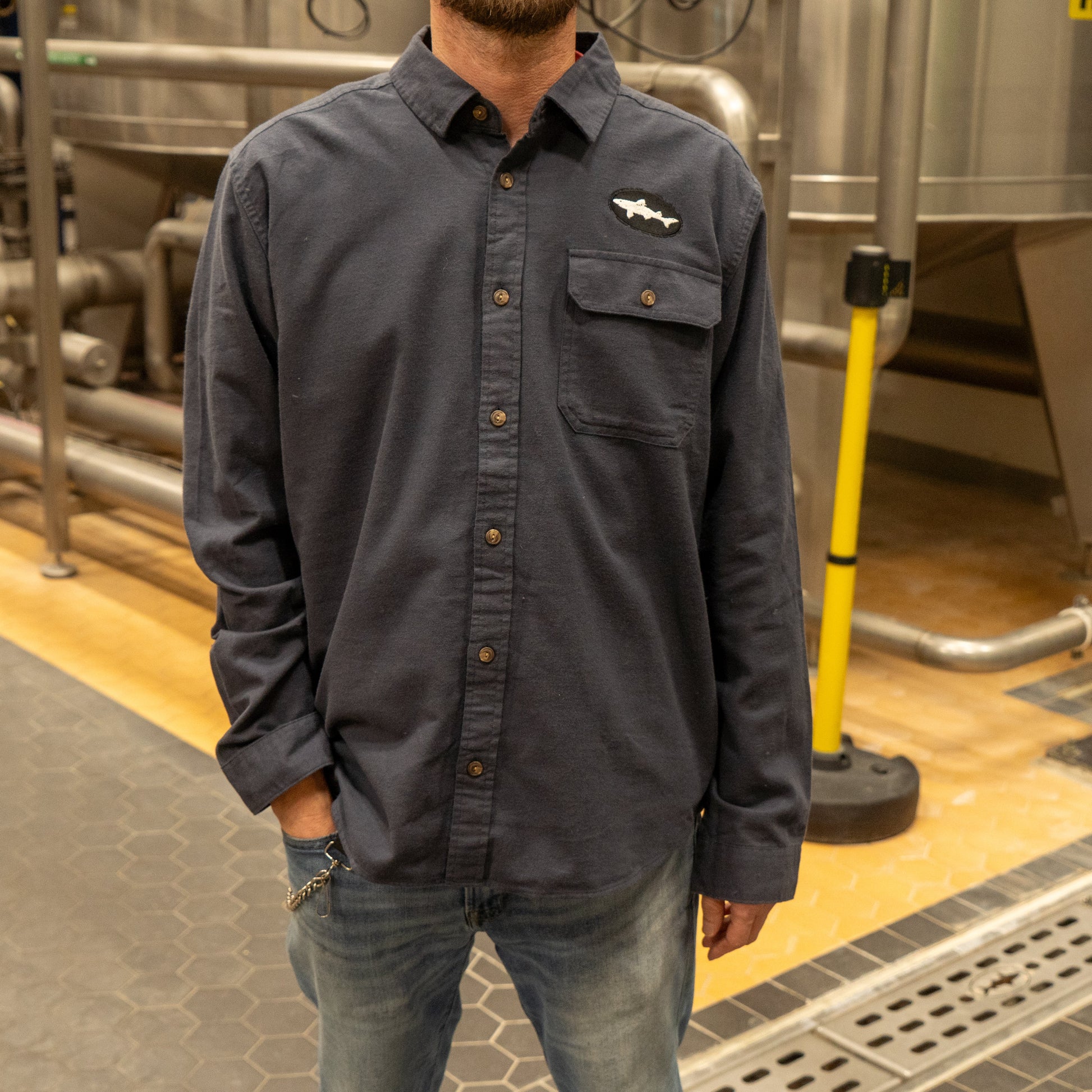 A man wearing a Patagonia M Ink Blue Light Weight Flannel Button Up with a chest pocket stands in an industrial setting with metal tanks and pipes in the background, his hand tucked into his pocket.