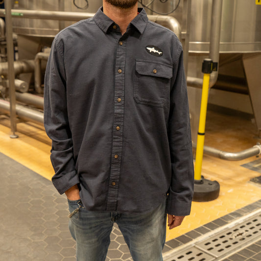 A man wearing a Patagonia M Ink Blue Light Weight Flannel Button Up with a chest pocket stands in an industrial setting with metal tanks and pipes in the background, his hand tucked into his pocket.