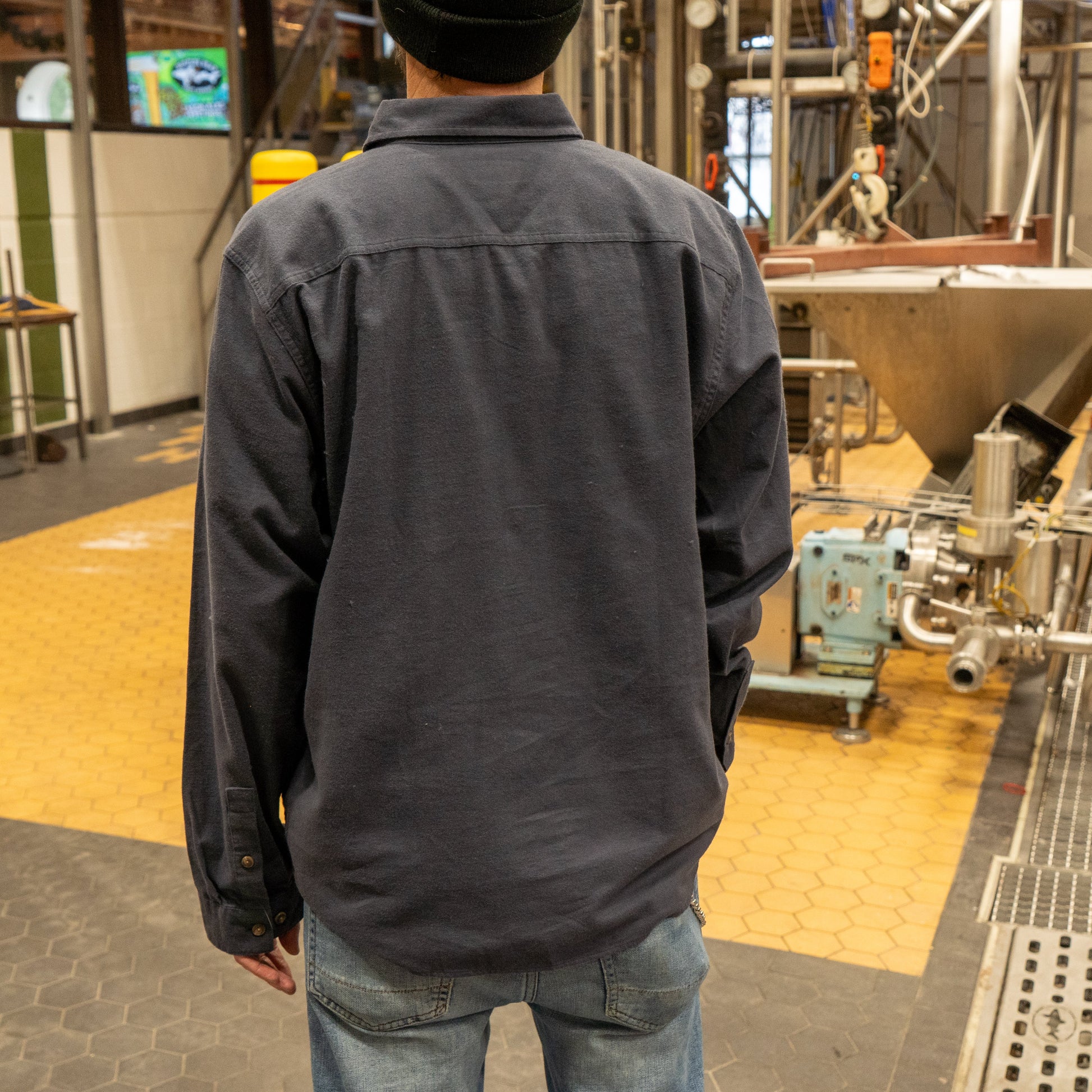 Wearing a Patagonia M Ink Blue Light Weight Flannel Button Up, jeans, and a black beanie, a person stands with their back to the camera in an industrial setting with yellow and black tiled floors and metal equipment.