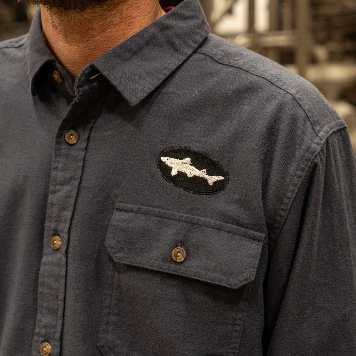 A person wears a Patagonia M Ink Blue Light Weight Flannel Button Up with a fish-shaped patch on the chest pocket. The patch is a black oval with a white fish outline. Only the shirt and part of the lower face are visible.