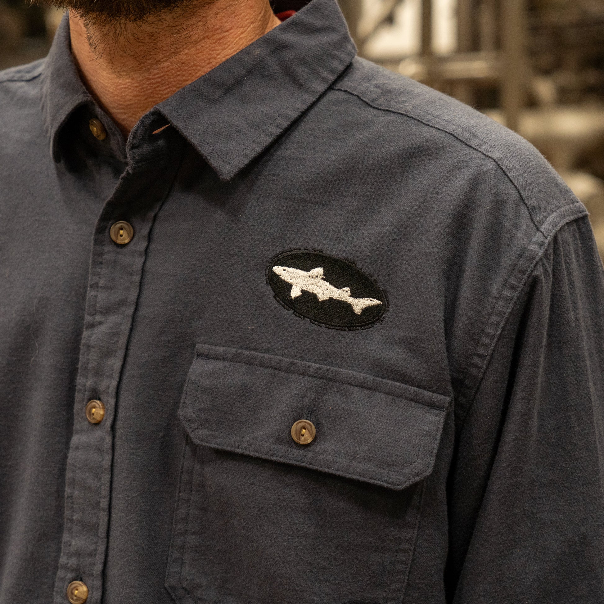 A person wears a Patagonia M Ink Blue Light Weight Flannel Button Up with a fish-shaped patch on the chest pocket. The patch is a black oval with a white fish outline. Only the shirt and part of the lower face are visible.