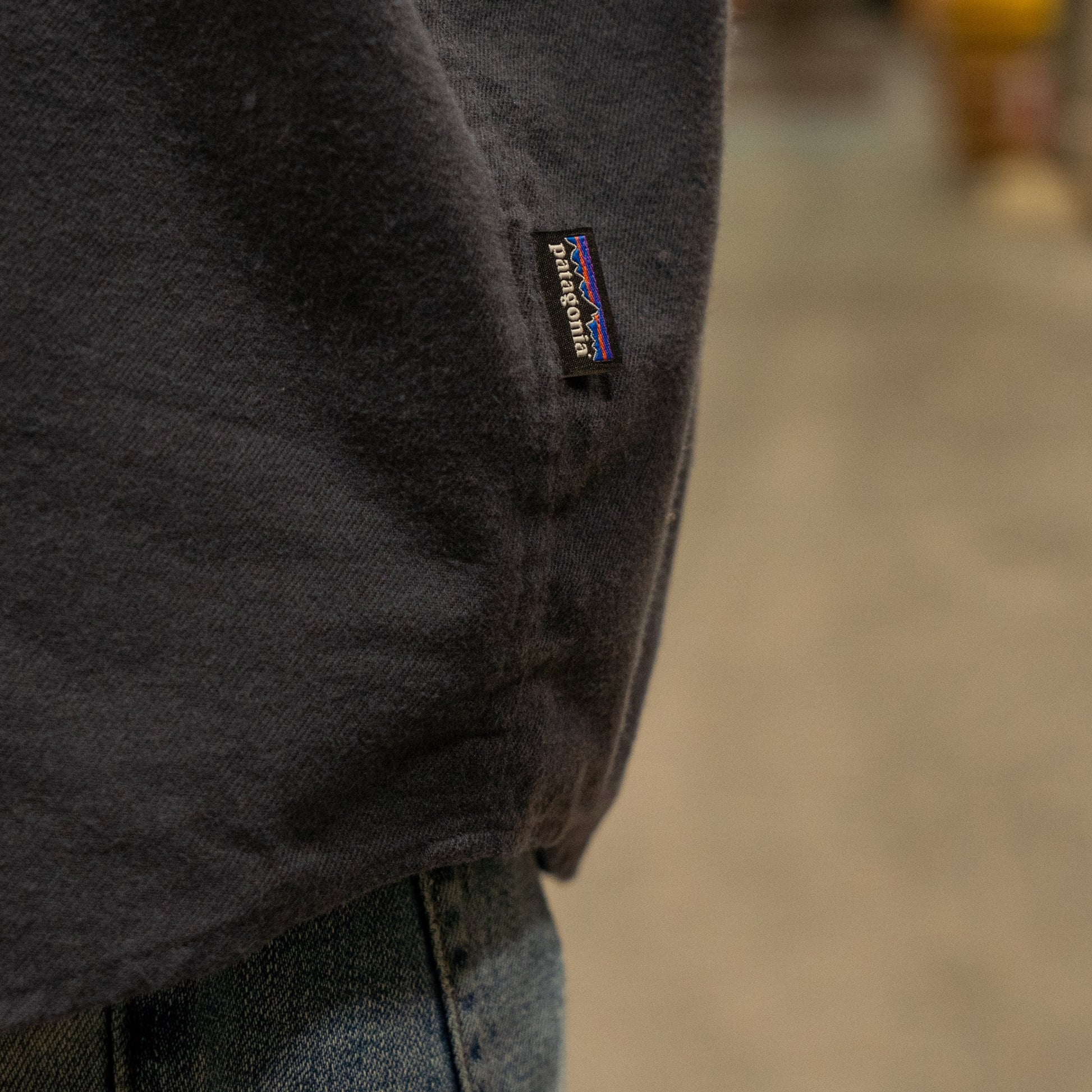 Close-up of a person wearing a Patagonia M Ink Blue Light Weight Flannel Button Up and blue jeans, highlighting the small black Patagonia tag on the shirt’s side seam. The background is blurred.