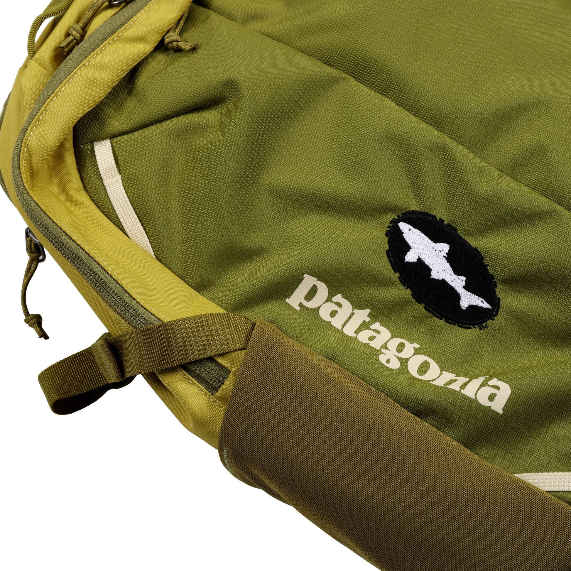 Close-up of the Patagonia Olive Refugio Day Pack 26L, showing the Patagonia logo and fish emblem. The 26L backpack features textured olive fabric, visible stitching, a strap, and a partially visible zipper.