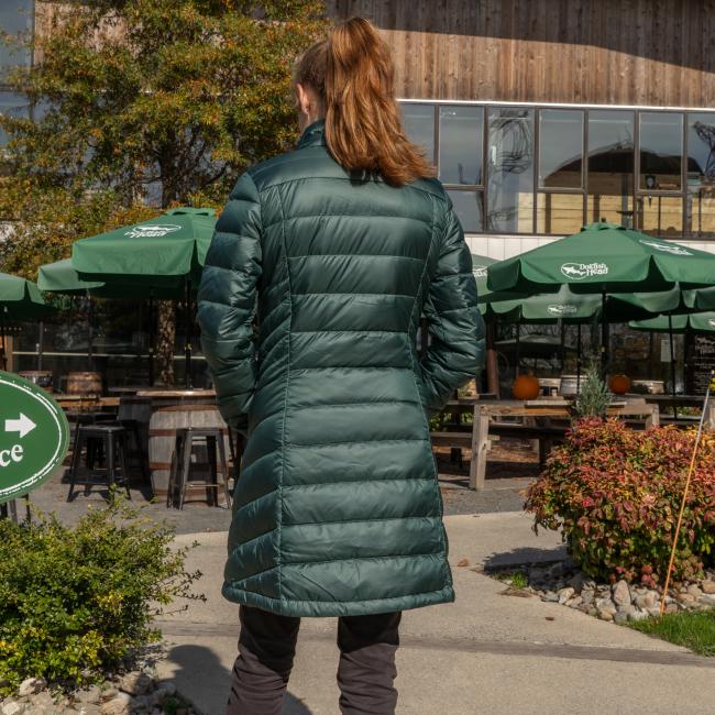 A woman with brown hair in a ponytail wears a Patagonia W Dark Green 3-in-1 Parka, standing outside with her back to the camera near green patio umbrellas and wooden tables at a café.