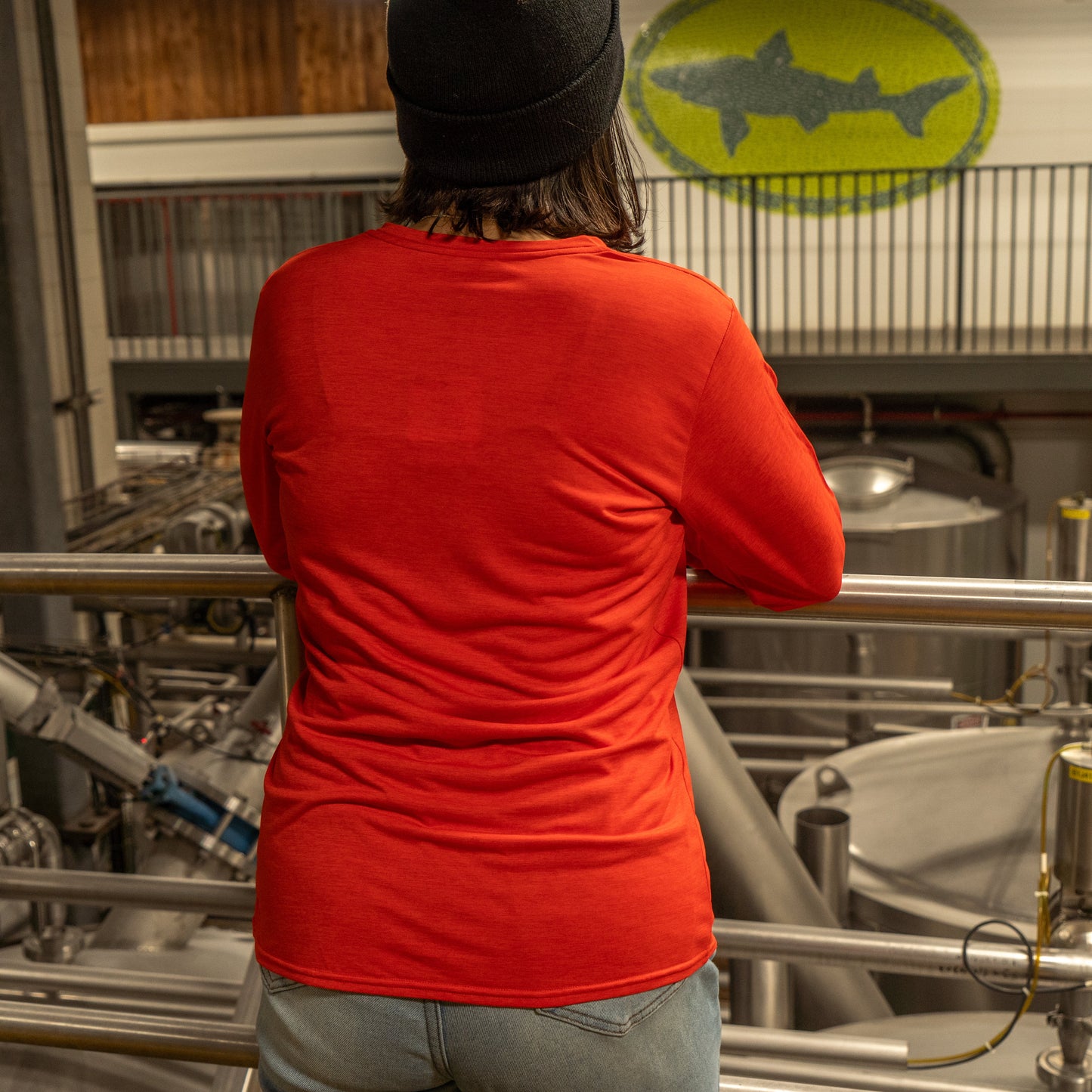 A person wearing a black beanie, jeans, and a Patagonia W Red Long Sleeve Runner stands with their back to the camera, overlooking brewery equipment. A large fish logo is visible on the wall in the background.