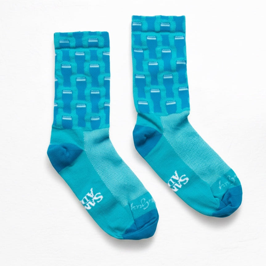 A pair of crew-length athletic socks in a gradient of turquoise and teal, featuring a repeating geometric pint glass pattern on the upper leg and white branding text on the toes.