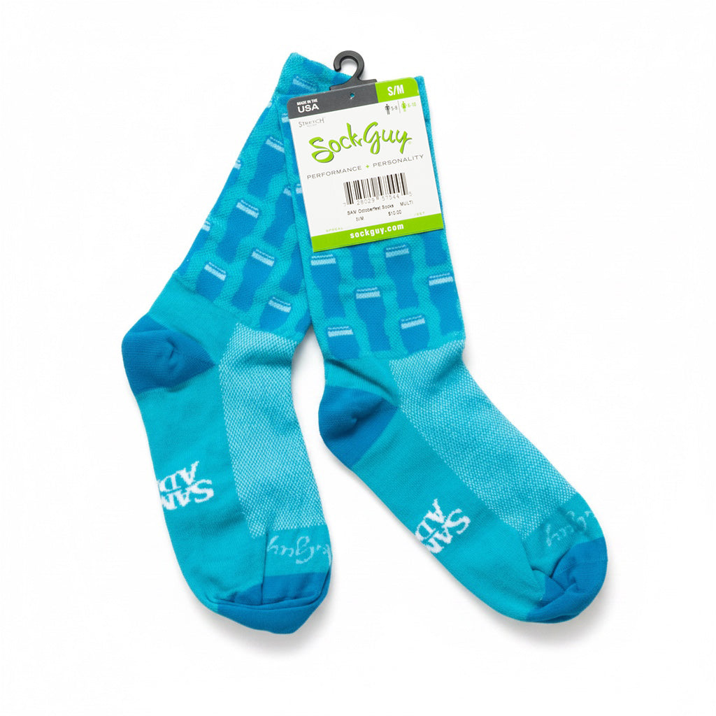 A pair of bright blue and turquoise crew socks with a geometric pint pattern, featuring the 'SockGuy' brand logo on the packaging and foot, displayed on a clean white background.