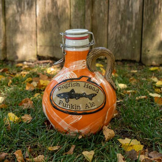 A ceramic jug labeled Mummy Punkin Growler 2 by Romanick Pottery, featuring an orange and white design, stands on grassy ground with yellow leaves and a wooden fence in the background.
