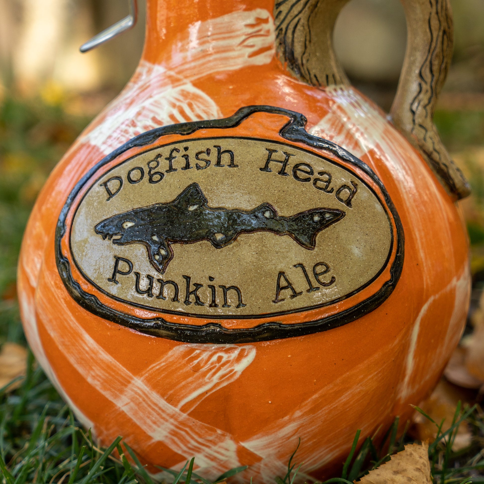 A close-up of an orange jug labeled Mummy Punkin Growler 2 from Romanick Pottery, shown outdoors on grass with some fallen leaves.