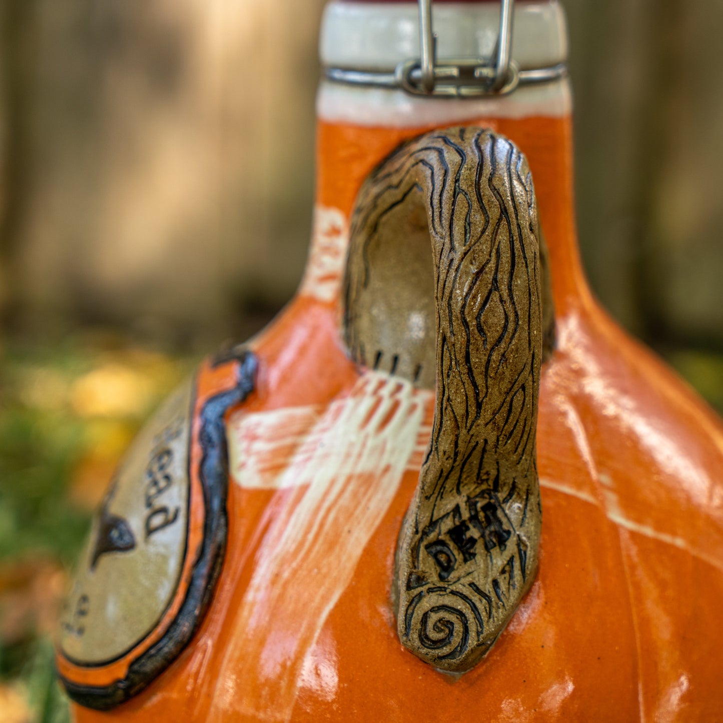 A close-up of the Mummy Punkin Growler 2 by Romanick Pottery, an orange ceramic jug with a sculpted animal ear handle, carved lines, and the word DEAF. The blurred background shows greenery and sunlight.