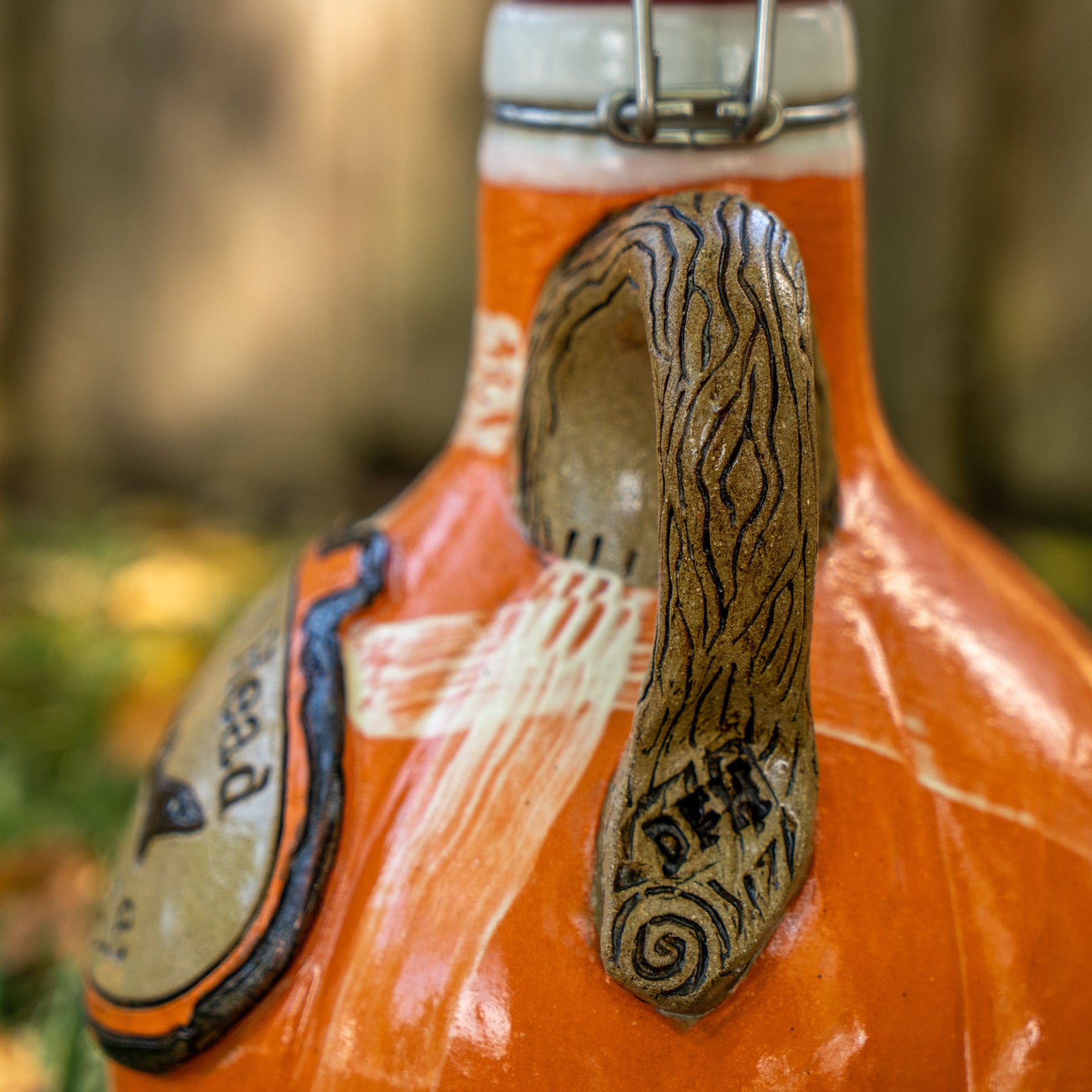 A close-up of the Mummy Punkin Growler 2 by Romanick Pottery, an orange ceramic jug with a sculpted animal ear handle, carved lines, and the word DEAF. The blurred background shows greenery and sunlight.