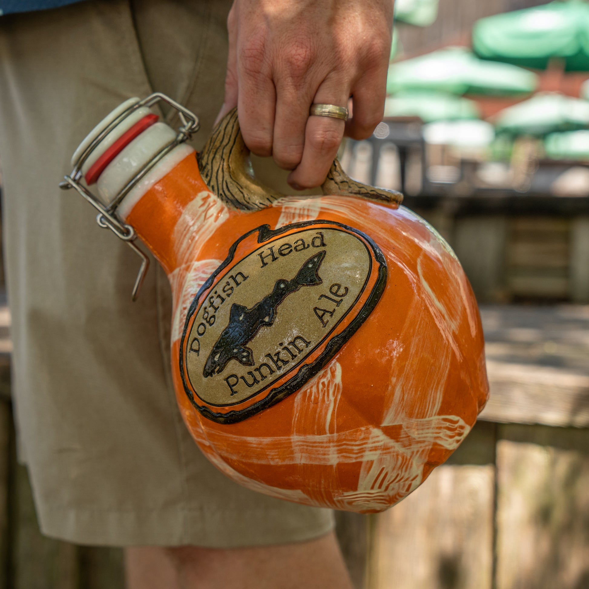 A person in shorts holds an orange Mummy Punkin Growler 2 by Romanick Pottery outdoors, with green umbrellas and wooden tables blurred in the background.