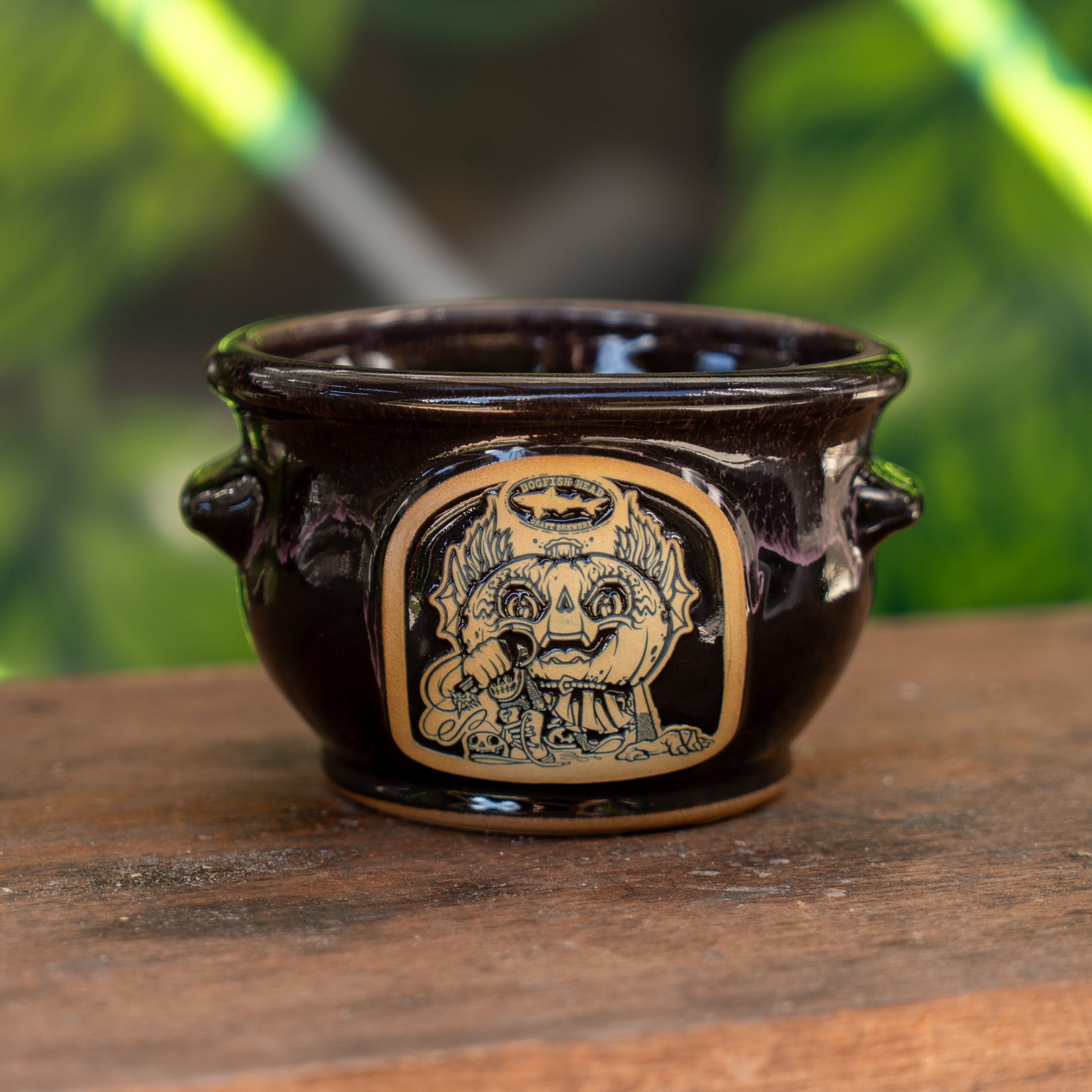 The Black Punkin Crock by Deneen Pottery, a dark glazed ceramic pot with side handles and a tan medallion featuring a whimsical crowned face, sits on wood, with blurred green foliage in the background.