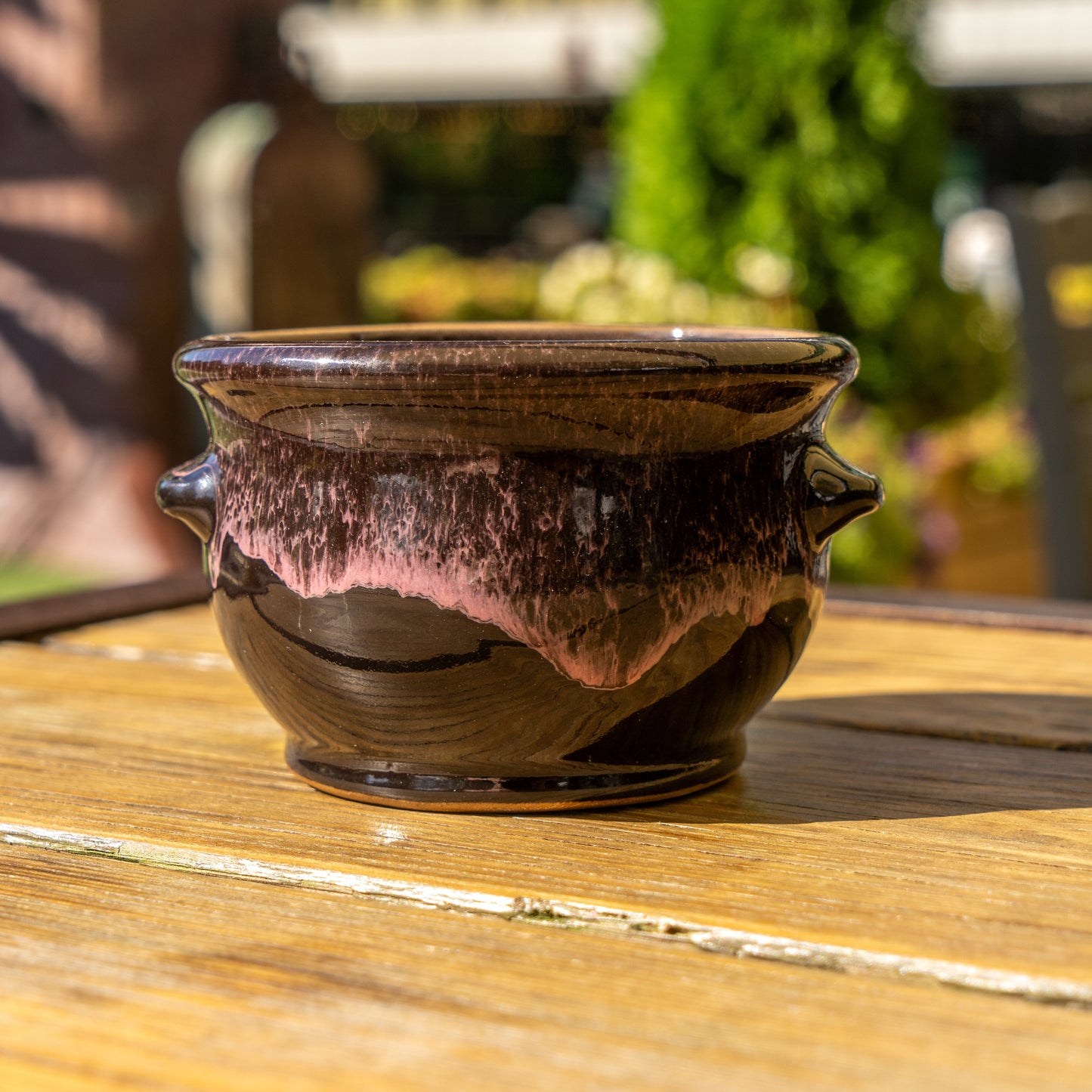 The Black Punkin Crock by Deneen Pottery, featuring a pinkish glossy glaze and small handles, sits on a wooden outdoor table in sunlight as part of a seasonal drop, with blurred greenery and buildings in the background.