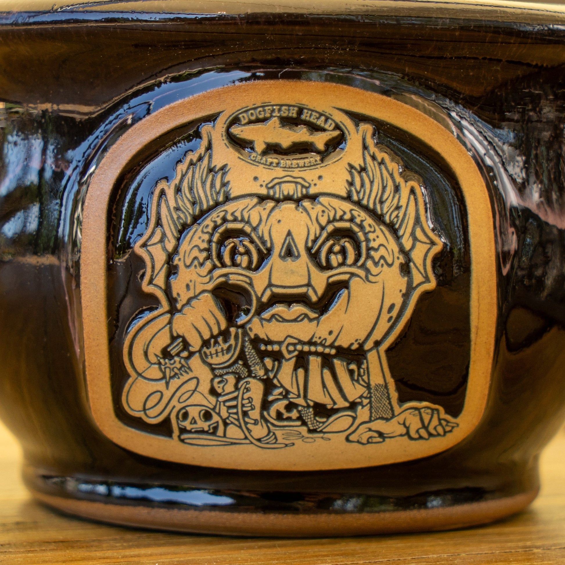 The Black Punkin Crock by Deneen Pottery is a ceramic mug featuring an engraved whimsical creature with large eyes, leafy ears, and a broad smile holding a hop cone—a playful design for this unique seasonal piece.