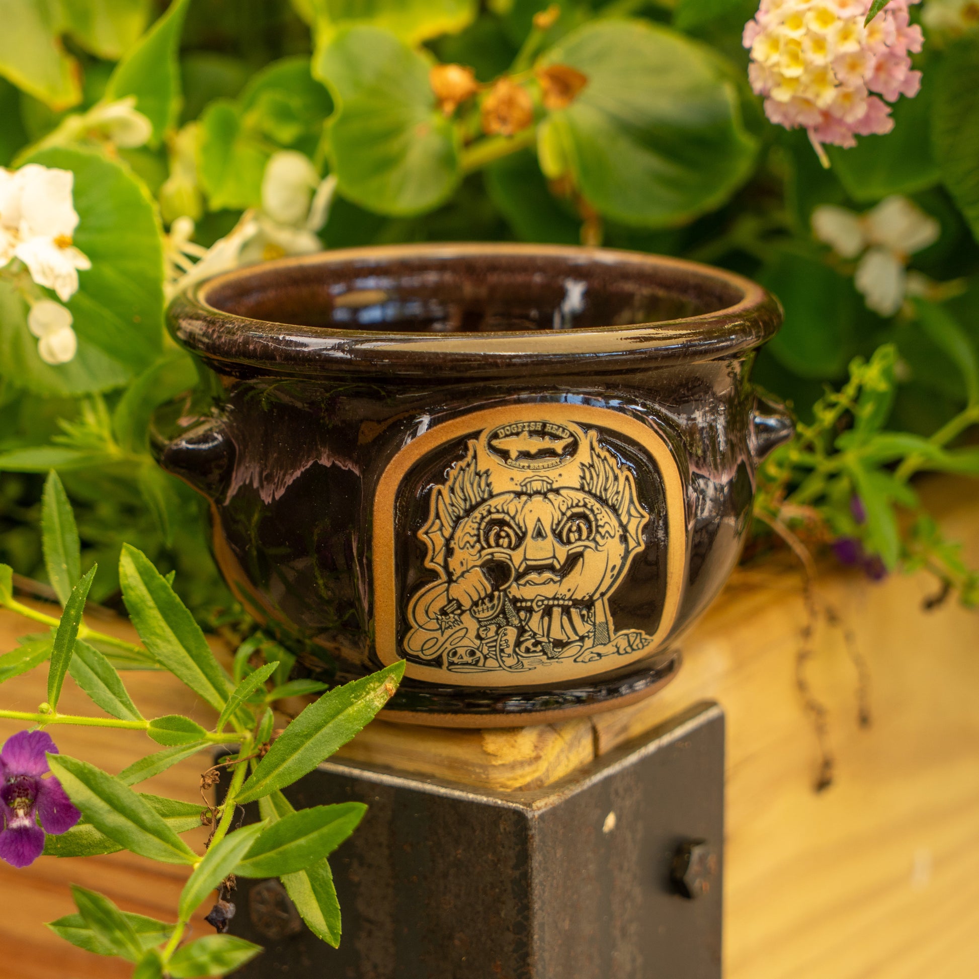The Black Punkin Crock by Deneen Pottery features a whimsical, hand-drawn cat face and sits on a wooden surface surrounded by green foliage and colorful flowers.