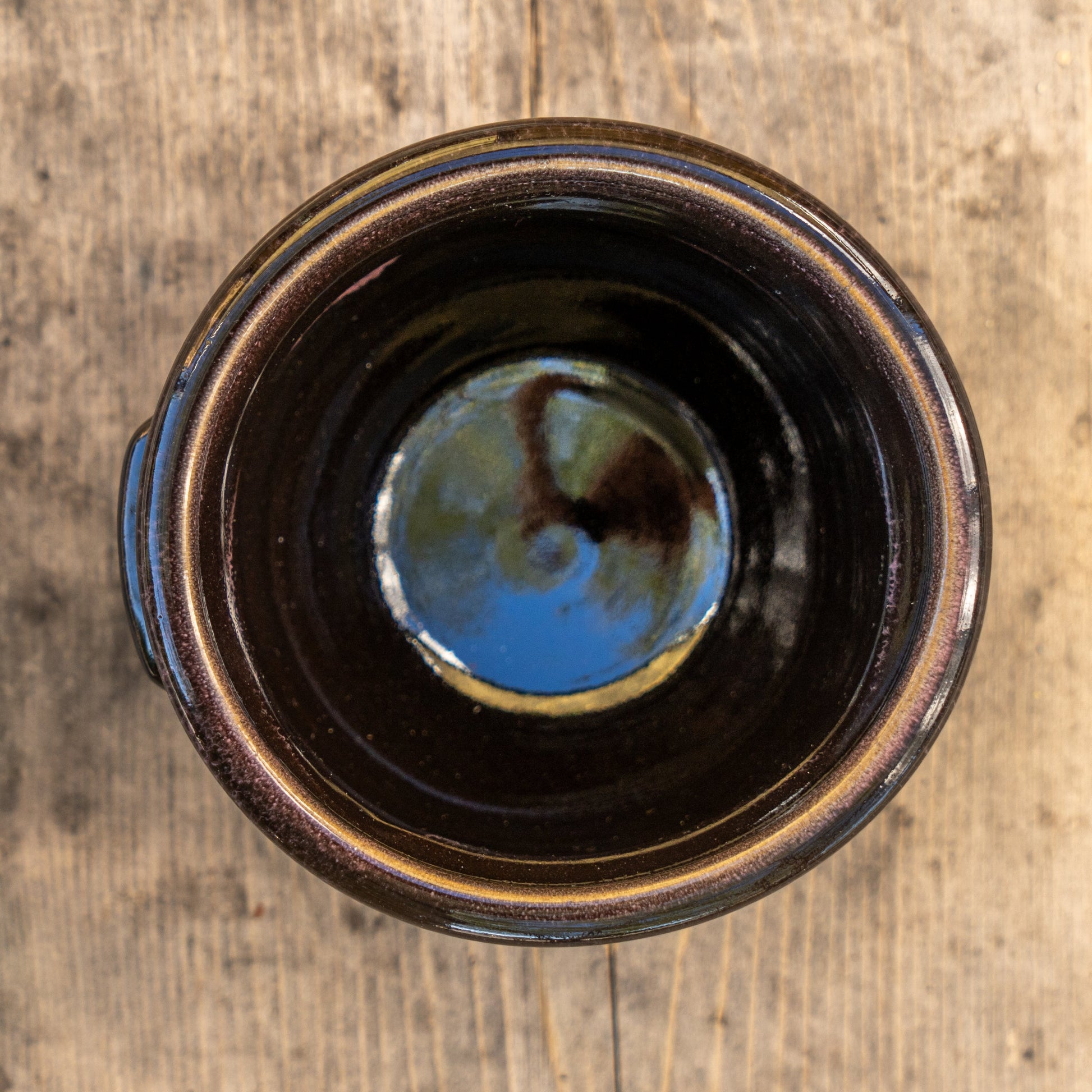 A top-down view of the Black Punkin Crock by Deneen Pottery reveals its reflective dark ceramic interior, echoing outdoor light and colors as it sits on a weathered wooden surface.