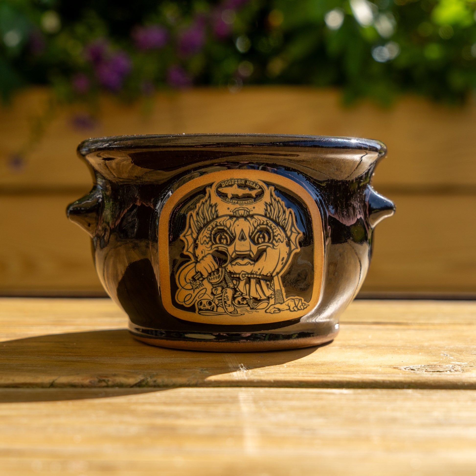 The Black Punkin Crock by Deneen Pottery is a black ceramic pot with two small handles and a stylized face design. It sits on a wooden surface, framed by greenery and purple flowers in the background.