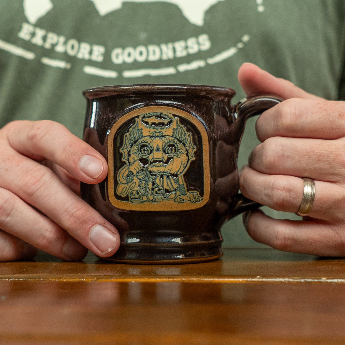 A person in a green EXPLORE GOODNESS shirt holds the Black Punkin Mug by Deneen Pottery, which features a pumpkin-headed figure hugging a smaller pumpkin-headed figure—ideal for seasonal drinkware.