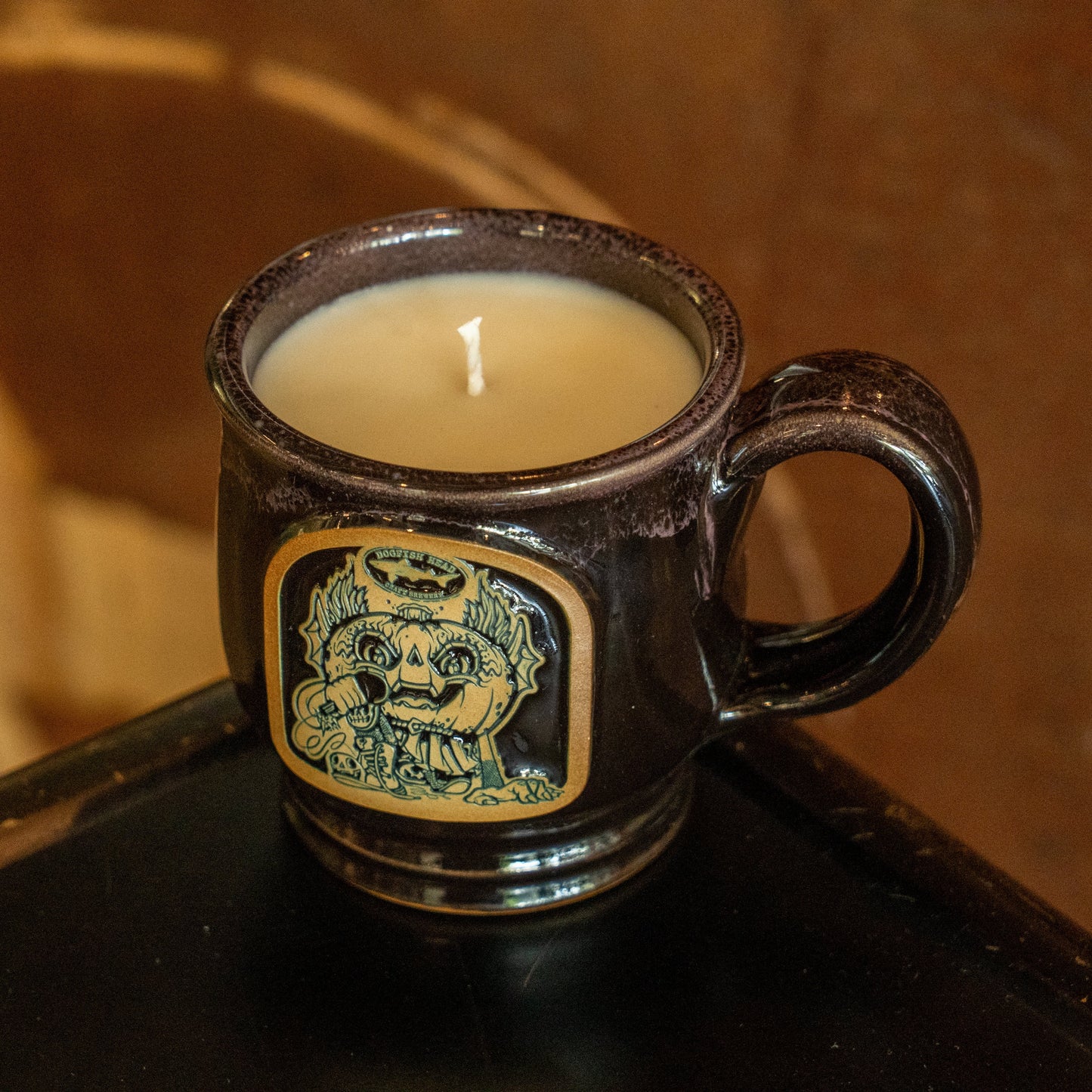 Experience the cozy fall apple crumble scent of the Black Punkin Mug Candle by Unwined Candles, LLC. This dark ceramic mug candle features a handle and a yellow and blue illustration of a grinning, fanged creature on the front. The stylish mug rests on a black surface against a blurred brown background.