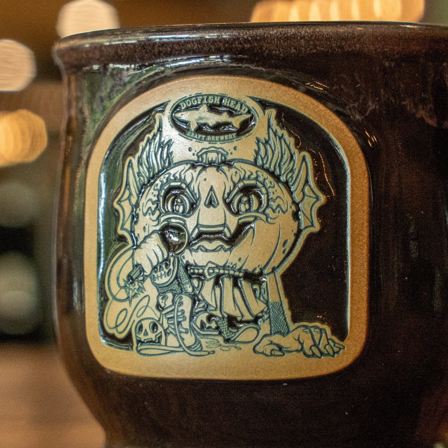 Introducing the Black Punkin Mug Candle by Unwined Candles, LLC—this unique candle features an illustration of a pumpkin-headed creature with horns, one hand holding a key and the other resting on a skull, with the Dogfish Head logo above the image. Infused with a cozy fall apple crumble scent, it’s perfect for enjoying your favorite autumn drink.