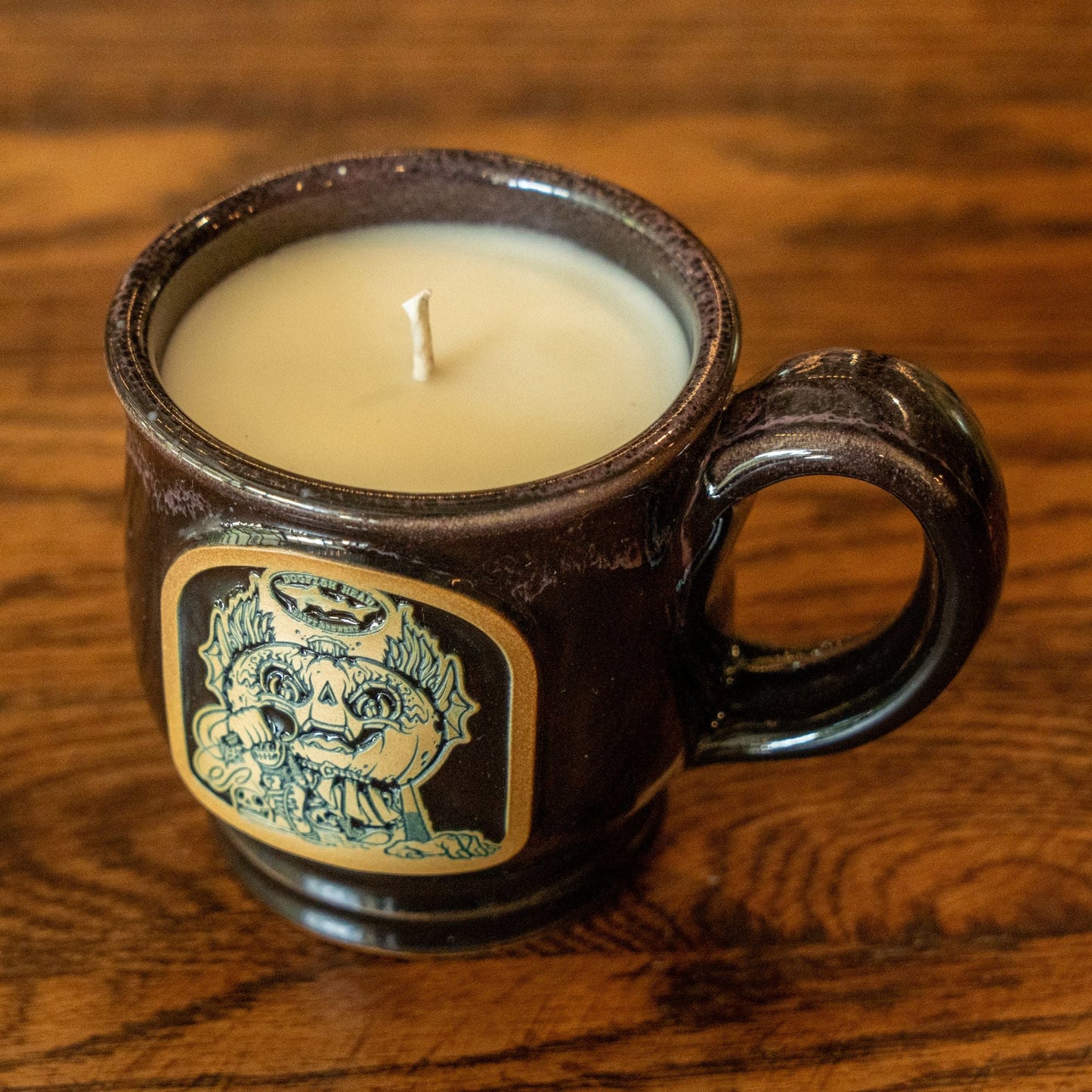 A Black Punkin Mug Candle by Unwined Candles, LLC with a fall apple crumble scent sits on a wooden surface. The mug features a decorative label with intricate artwork on the front, evoking the warmth of an autumn drink.
