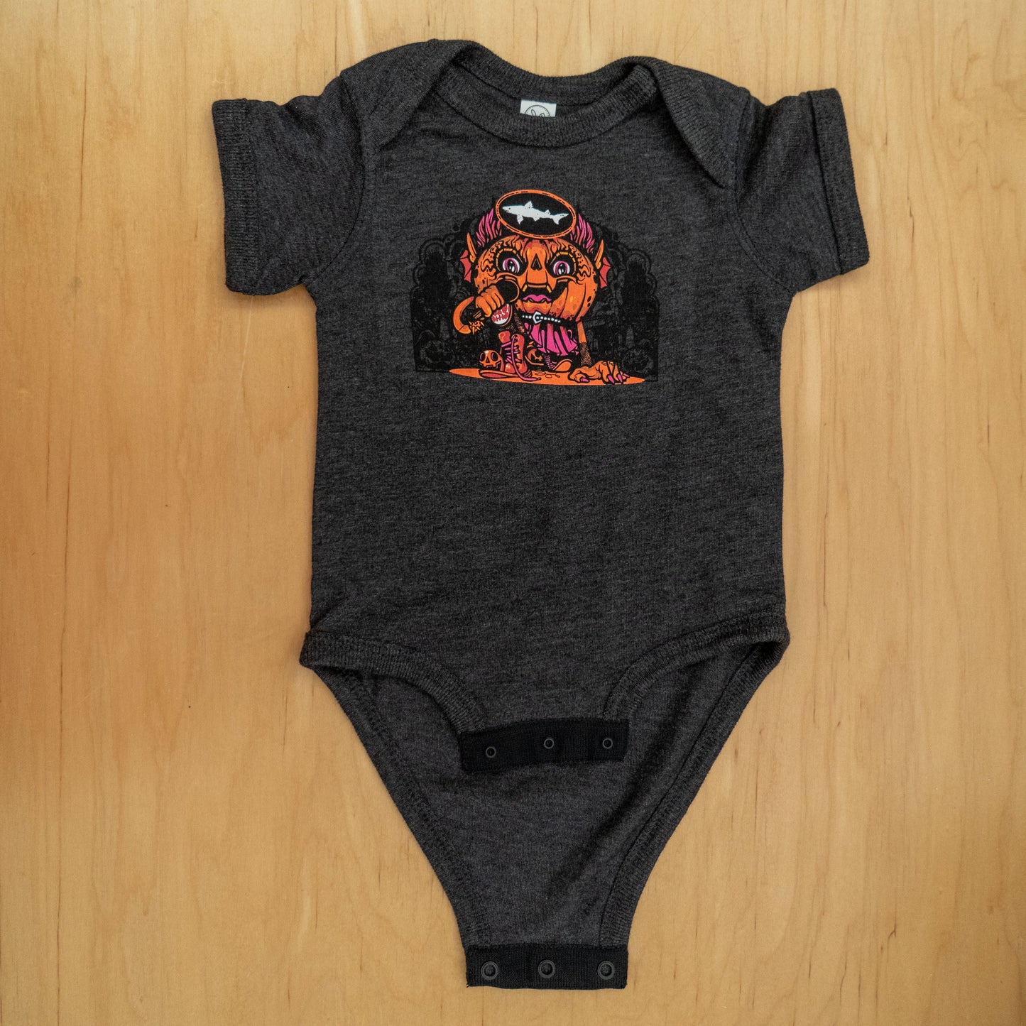 The Punkin Onesie by GO WEST T-SHIRT COMPANY is a dark gray baby bodysuit featuring Drew Millward’s colorful cartoon of an orange monster with a drumstick, bones, and bat bib, perfect for fall. Laid flat on a wooden surface.