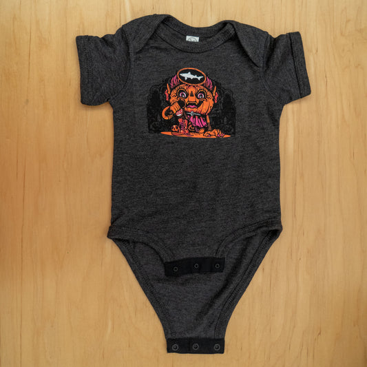 The Punkin Onesie by GO WEST T-SHIRT COMPANY is a dark gray baby bodysuit featuring Drew Millward’s colorful cartoon of an orange monster with a drumstick, bones, and bat bib, perfect for fall. Laid flat on a wooden surface.