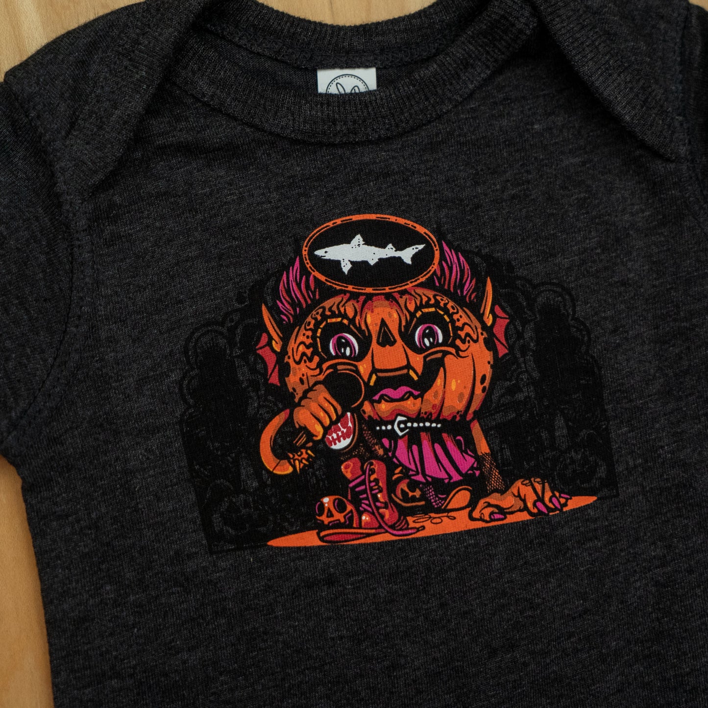 The Punkin Onesie by GO WEST T-SHIRT COMPANY is a dark gray fall outfit with a colorful cartoon pumpkin holding a microphone, wearing a pink cape and crown, inspired by Drew Millward, plus fun fish, bones, and tentacles details.
