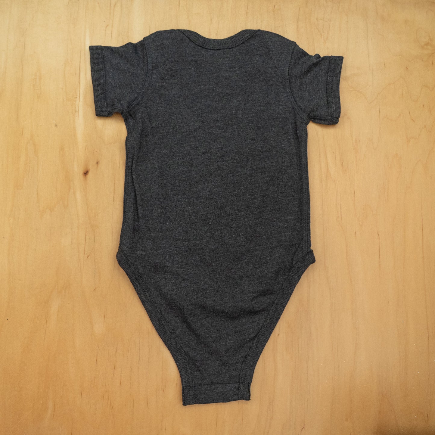 The GO WEST T-SHIRT COMPANY Punkin Onesie is a dark gray, short-sleeved fall baby outfit, pictured laid flat on a light wooden surface with the back view—ideal for seasonal wear.
