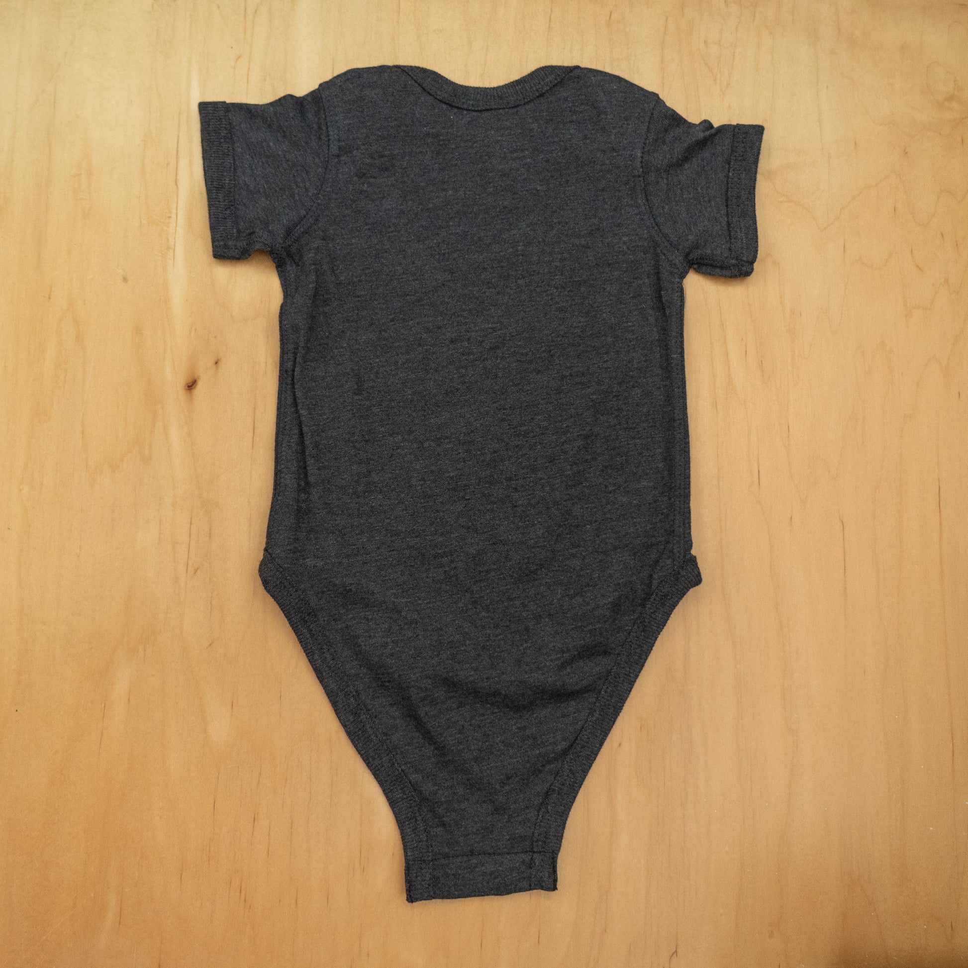 The GO WEST T-SHIRT COMPANY Punkin Onesie is a dark gray, short-sleeved fall baby outfit, pictured laid flat on a light wooden surface with the back view—ideal for seasonal wear.