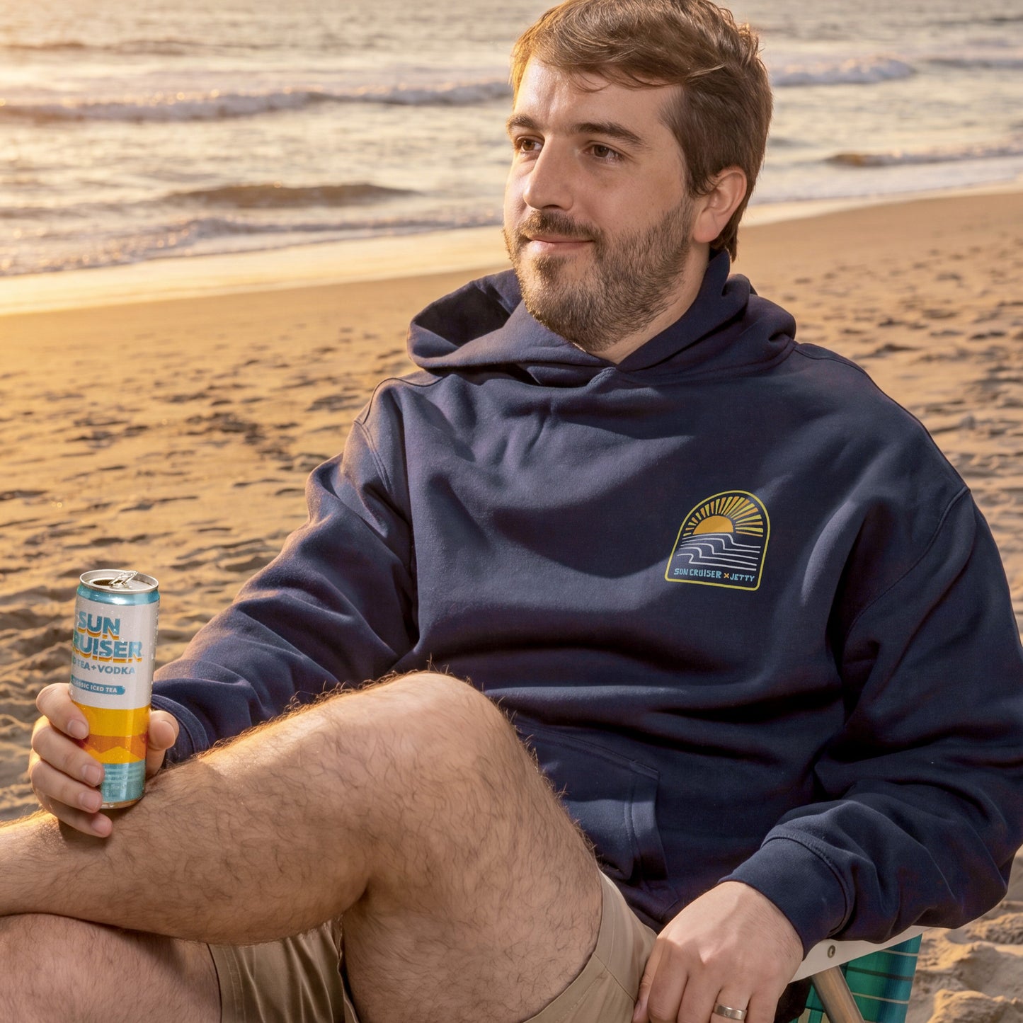Man sitting on a beach holding a can, wearing a navy hoodie with a logo.