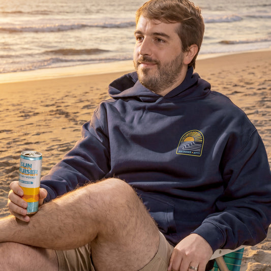 Man sitting on a beach holding a can, wearing a navy hoodie with a logo.