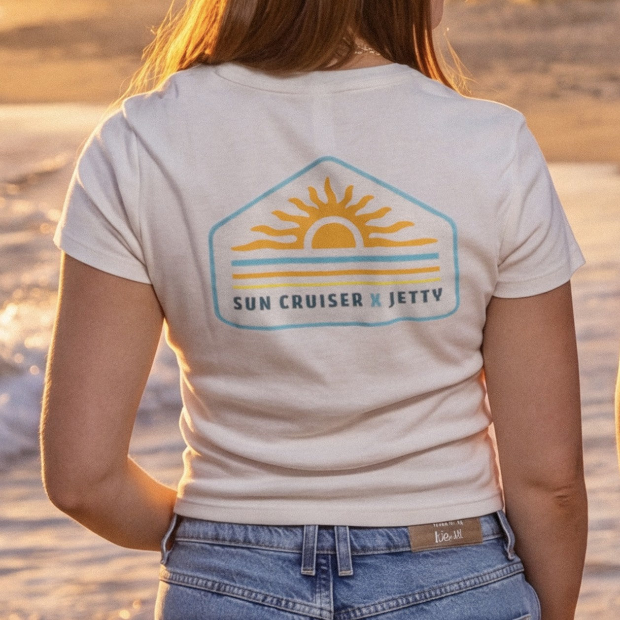 person wearing a sleeve cream tee shirt with Sun cruiser x Jetty logo on back on a beach. 
