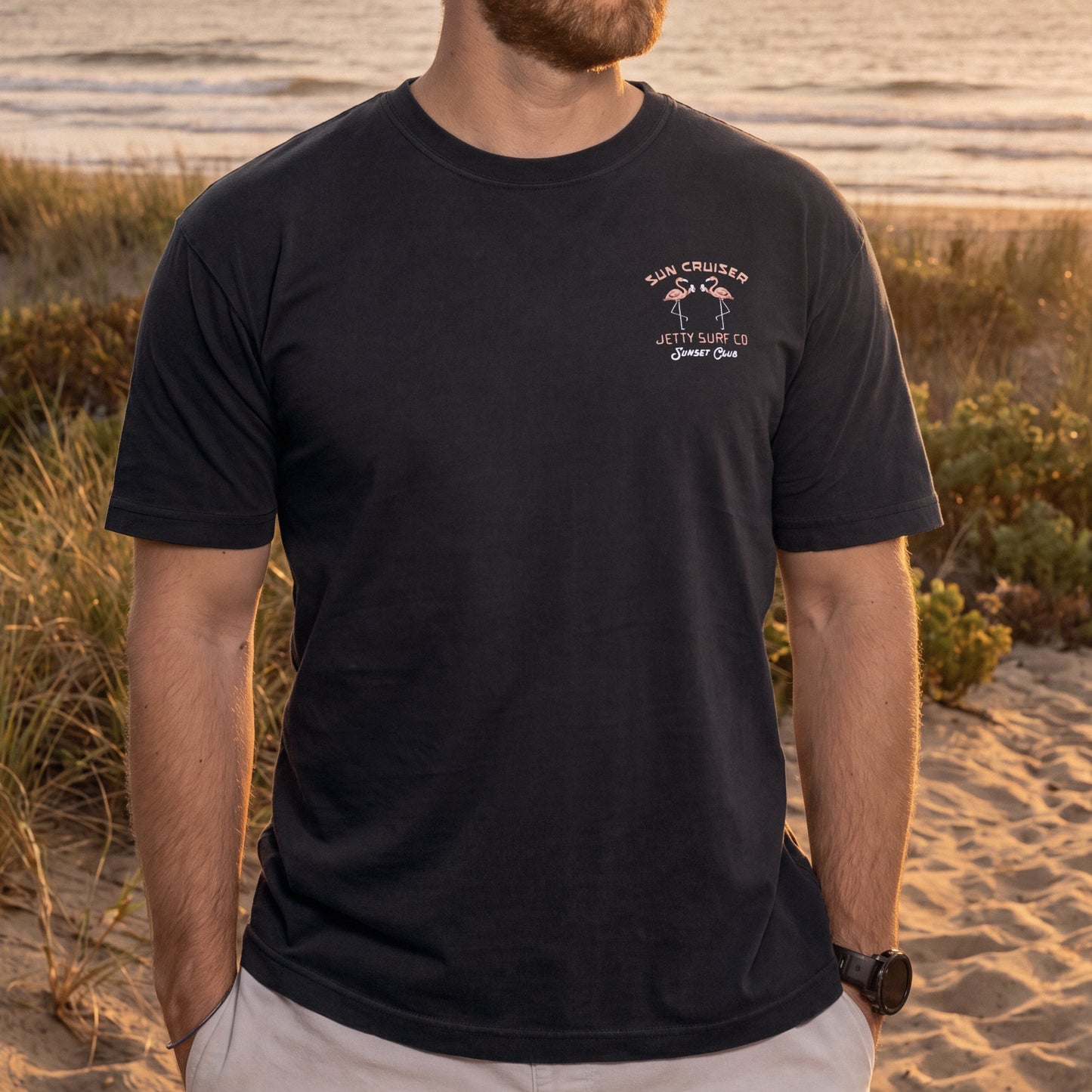 A man wearing a black crewneck t-shirt with a small white graphic on the left chest that reads 'Sun Cruiser' and 'Jetty Surf Co', standing on a sandy beach with dunes and water in the background.