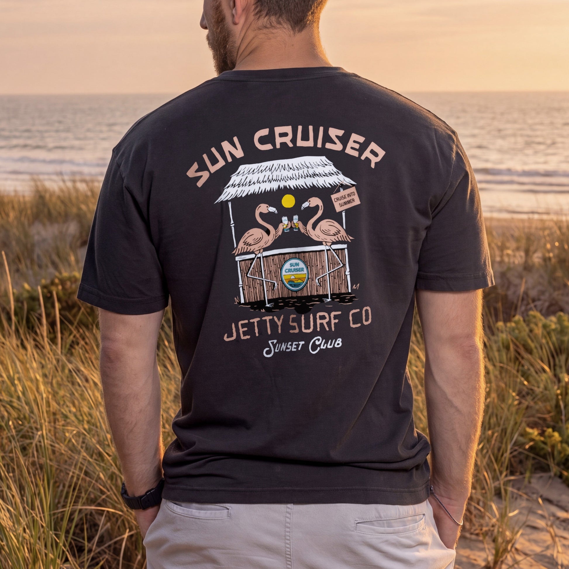 A man standing on a beach dune at sunset, viewed from behind, wearing a dark charcoal t-shirt with a large graphic print on the back featuring flamingos, a beach cart, and the text 'SUN CRUISER' and 'JETTY SURF CO Sunset Club'.