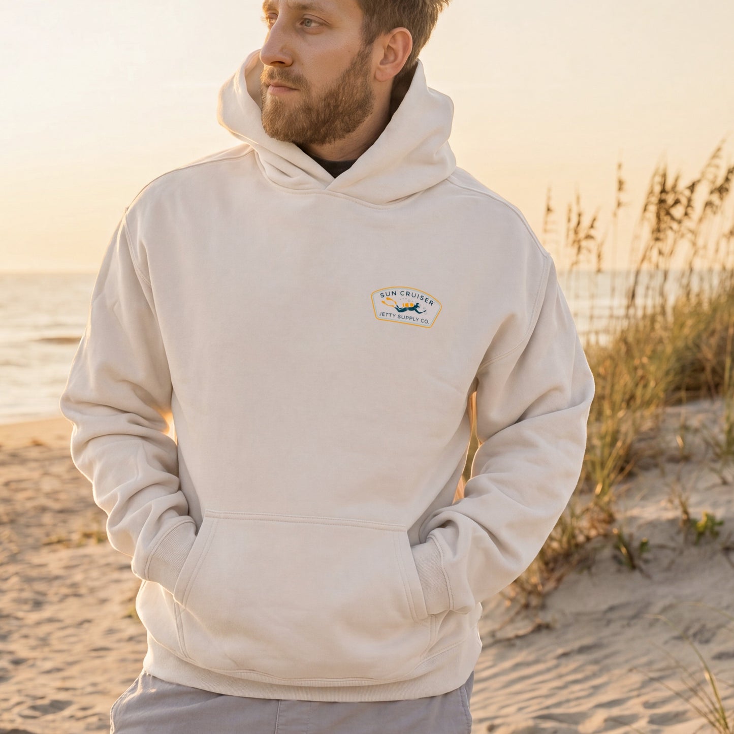 Man wearing a beige hoodie with a logo on a beach at sunset