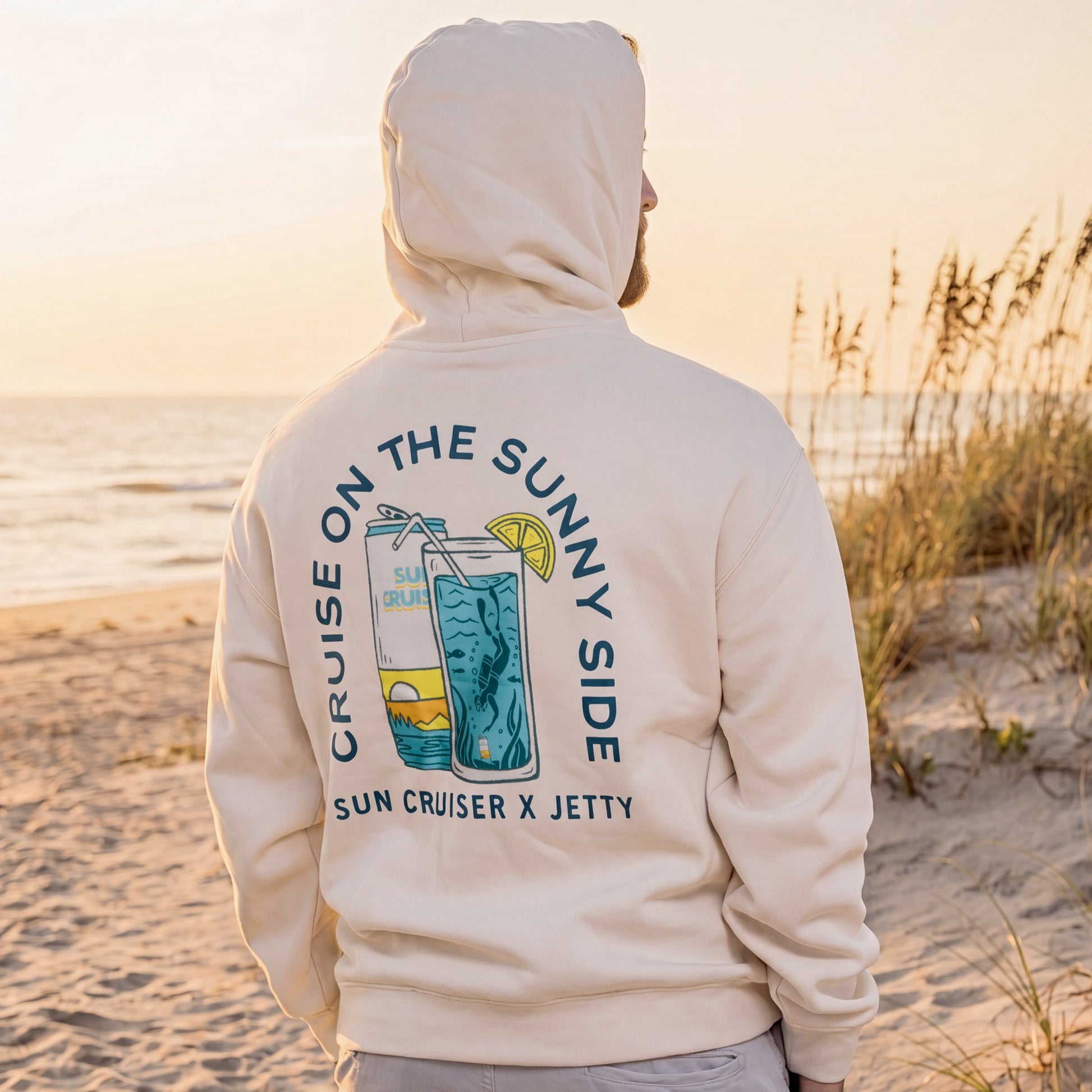 Person wearing a hoodie with a beach-themed design on a sandy beach at sunset.