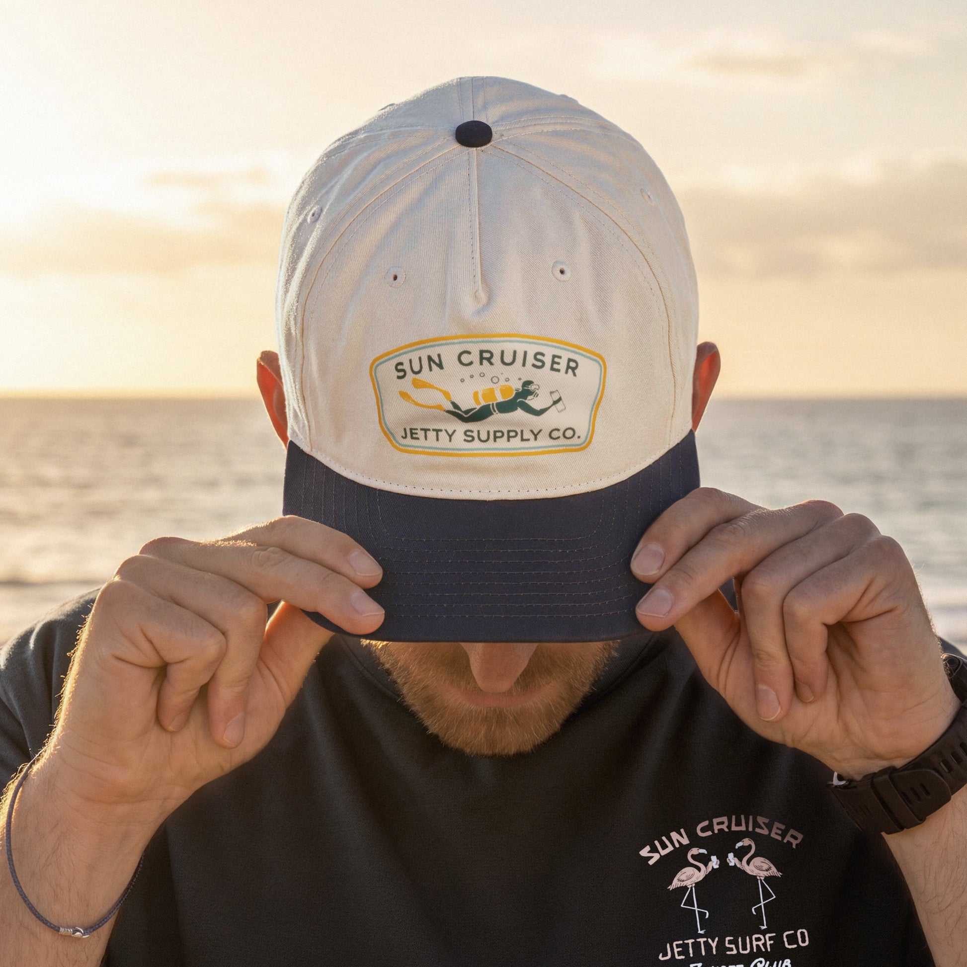 A man wearing a white and navy blue baseball cap with a 'Sun Cruiser Jetty Supply Co.' patch on the front, holding the brim down over his eyes.