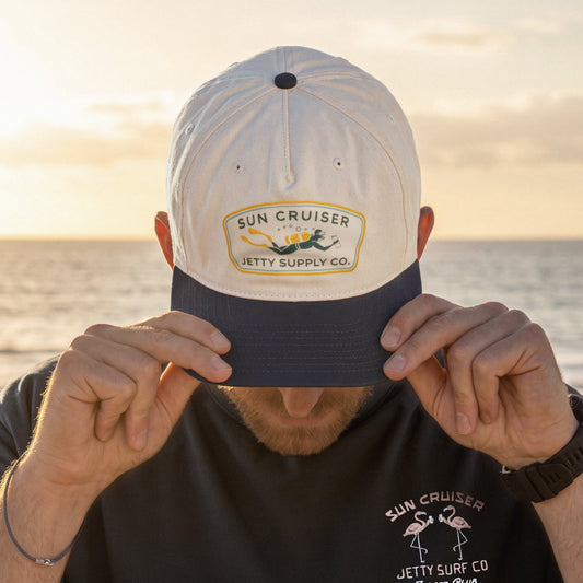 A man wearing a white and navy blue baseball cap with a 'Sun Cruiser Jetty Supply Co.' patch on the front, holding the brim down over his eyes.