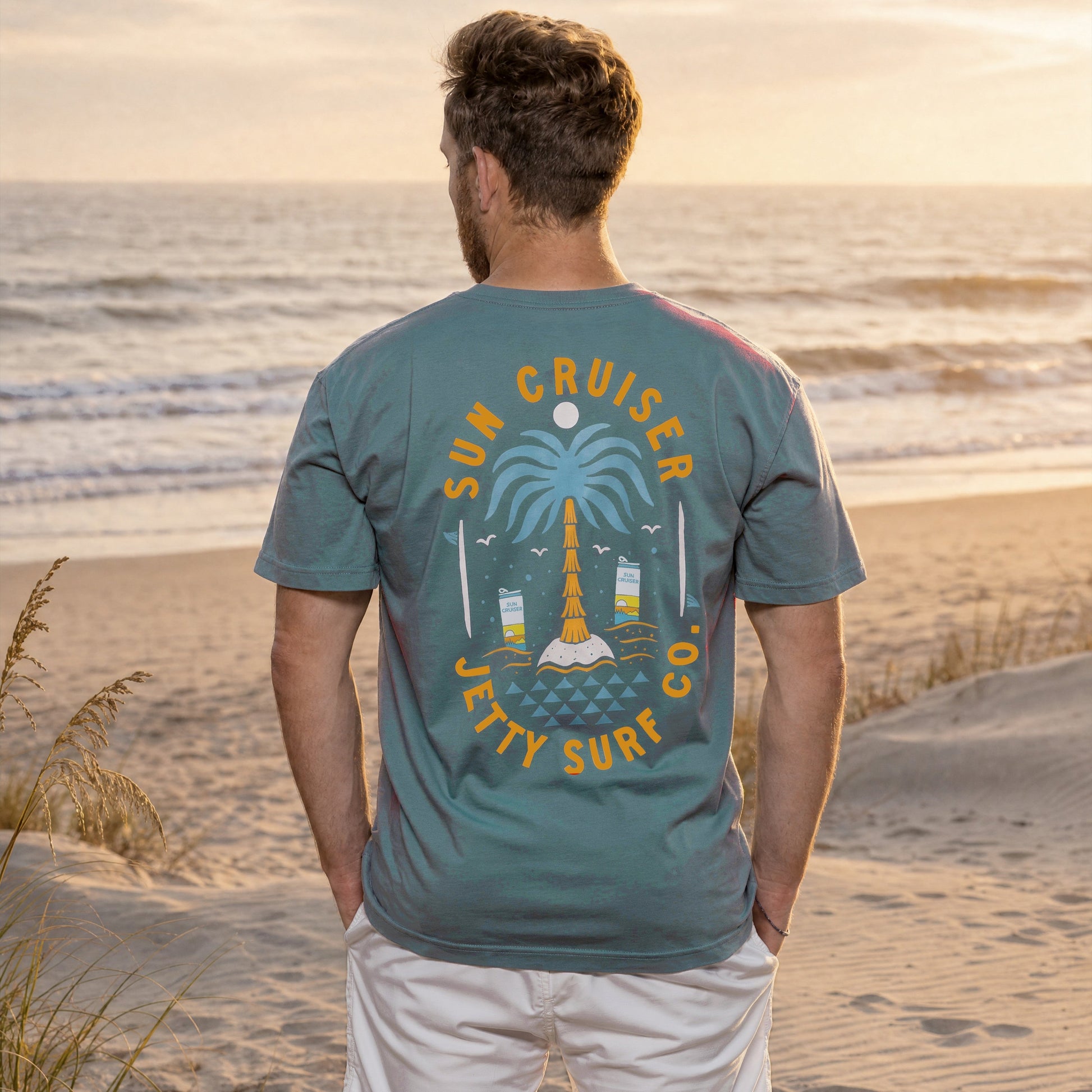 Blue short sleeve tee on person with large Sun Cruiser Jetty surf co palm tree graphic on back. 