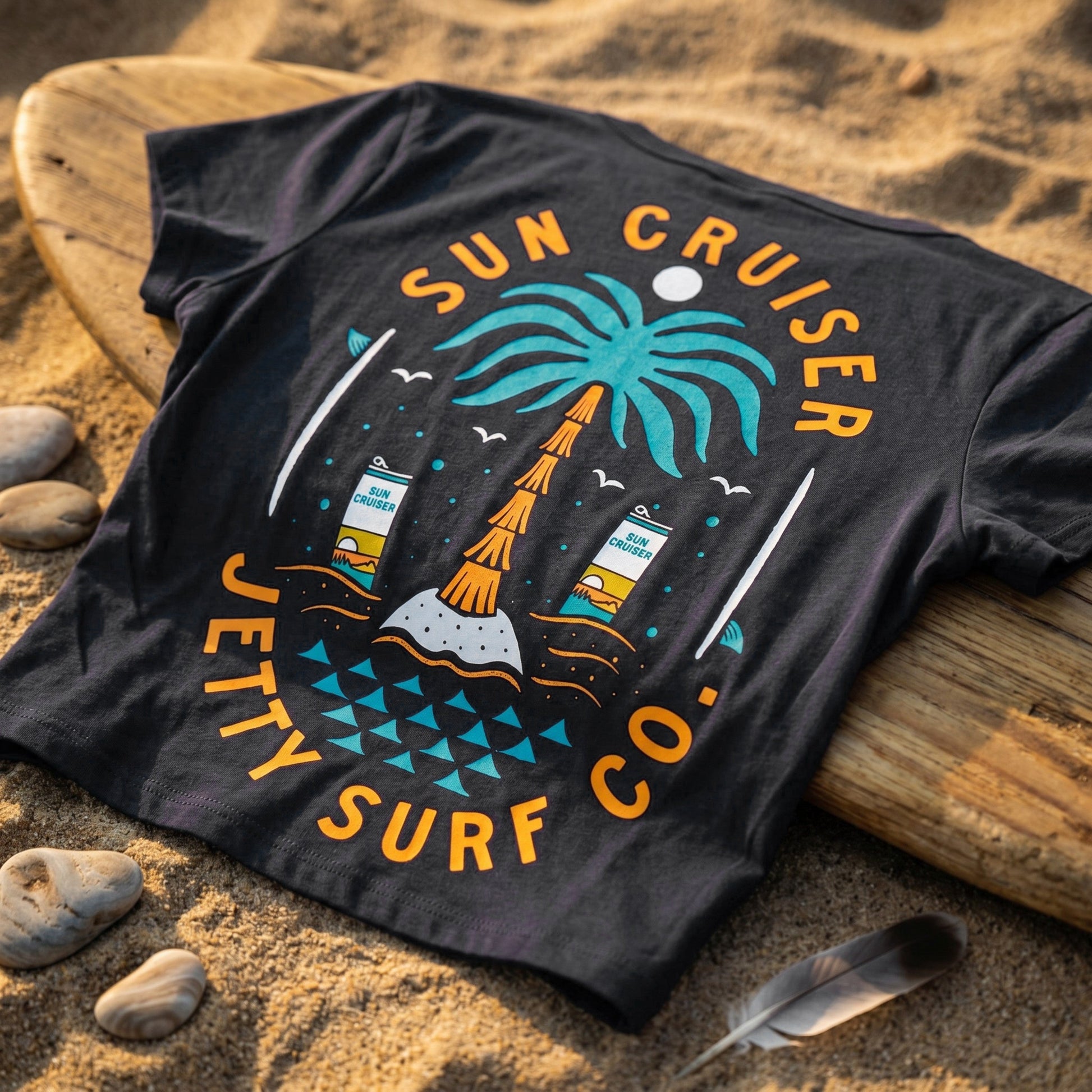 A black graphic t-shirt laid flat on a sandy beach next to a wooden surfboard, featuring a colorful tropical beach scene with the text 'SUN CRUISER' and 'JETTY SURF CO.'.