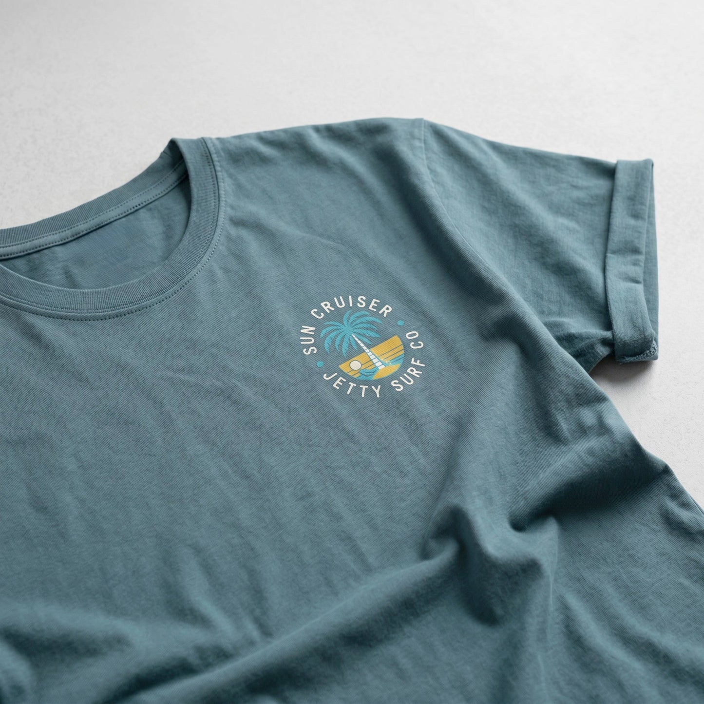 A slate blue crewneck t-shirt featuring a circular 'Sun Cruiser, Jetty Surf Co.' graphic on the left chest with a palm tree and yellow sunset illustration.